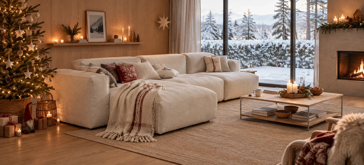 A large cream-colored modular sectional sofa in a cozy, warmly lit living room decorated for Christmas, with a fireplace, a lit tree, and a large window showing a snowy outdoor view.