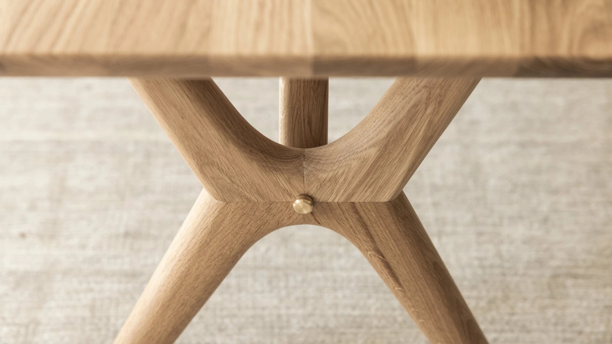 Close-up of a light oak wood dining table base with a sculptural X-shaped leg design and a small brass center rivet, set on a neutral textured rug.