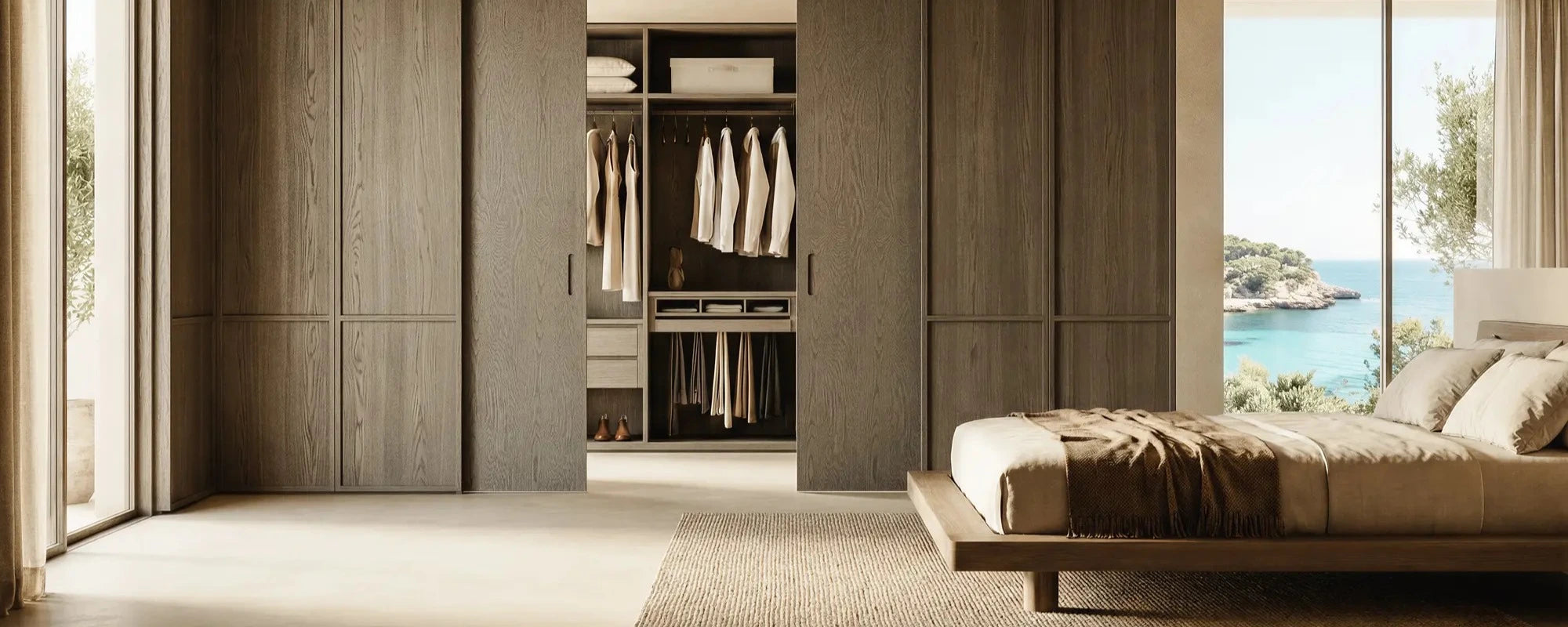 A modern bedroom interior featuring a large built-in wooden wardrobe with sliding doors and open shelving, next to a low platform bed with neutral bedding and a large window overlooking the ocean.