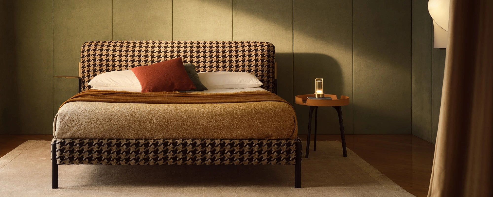A bed with a brown and beige houndstooth patterned frame, dressed in beige and brown bedding with a rust-colored accent pillow, set in a dimly lit bedroom with a small wooden side table.